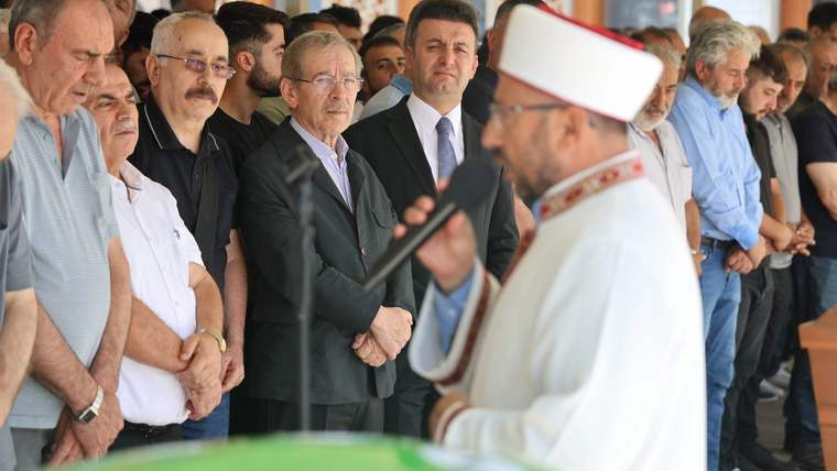 Beyaz başlıklı bir din görevlisi, önünde yeşil örtülü bir bölüm bulunan salonda sıralanan insanların yanında duruyor.
