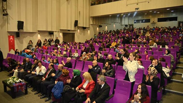 Mor koltuklu salonda çocuklar ve yetişkinler otururken bazı kişiler alkışlıyor; sol tarafta Türk bayrağı görülüyor.