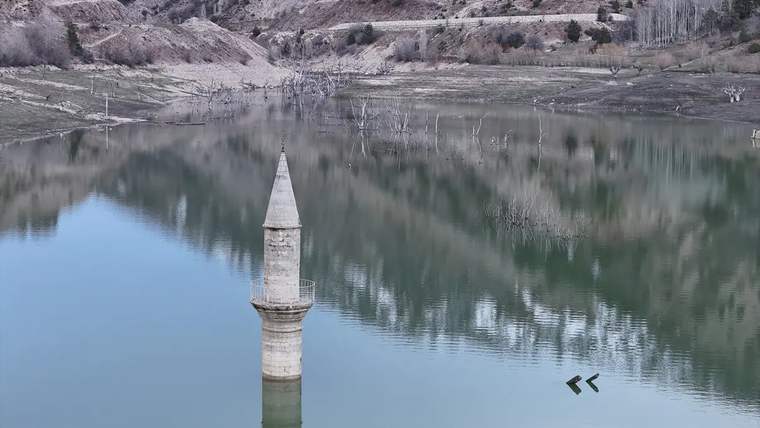 Sular altında kalan minarenin yalnızca üst bölümü, durgun göl ve çıplak yamaçların önünde görülüyor.