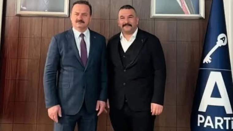 Ahşap panelli duvar önünde takım elbiseli iki erkek ve yanında Anahtar Parti logolu bayrak görülüyor.