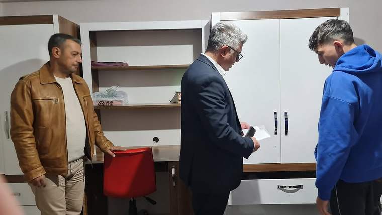 Bir odada üç erkek bulunuyor; ortadaki kişi elindeki kâğıtlara bakarken diğer ikisi onu izliyor.