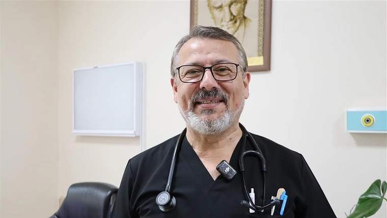 Siyah sağlık kıyafeti ve stetoskop takan, gözlüklü, sakallı bir kişi odada kameraya bakıyor.
