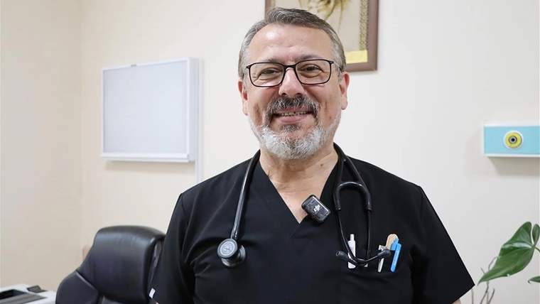 Siyah sağlık kıyafeti ve stetoskop takan, gözlüklü, sakallı bir kişi odada kameraya bakıyor.
