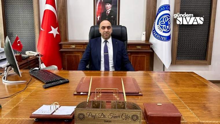Bir masa arkasında oturan bir kişi, masanın üzerinde dosyalar ve bir isimlik var.