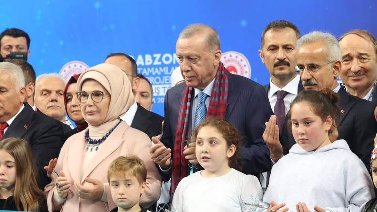 Mavi arka plan önünde, bir grup yetişkin ve çocuğun arasında duran erkek, mavi kurdelenin arkasında görülüyor.