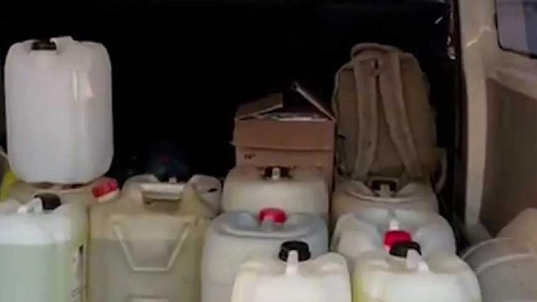 Karanlık bir araç bölümünde çeşitli boyutlarda beyaz plastik bidonlar ve bir sırt çantası görülüyor.