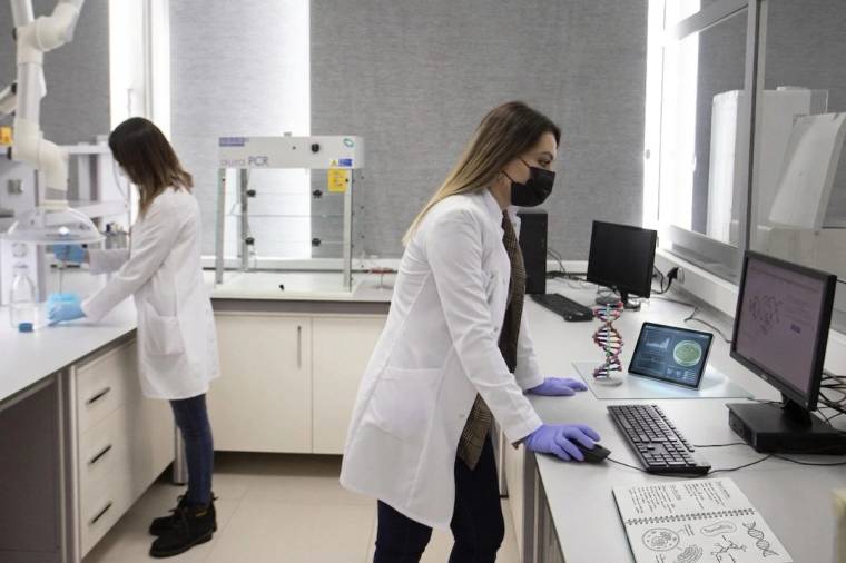 Laboratuvarda iki beyaz önlüklü kadın çalışıyor; öndeki maskeli kadın bilgisayar ve deney ekipmanlarının yanında.