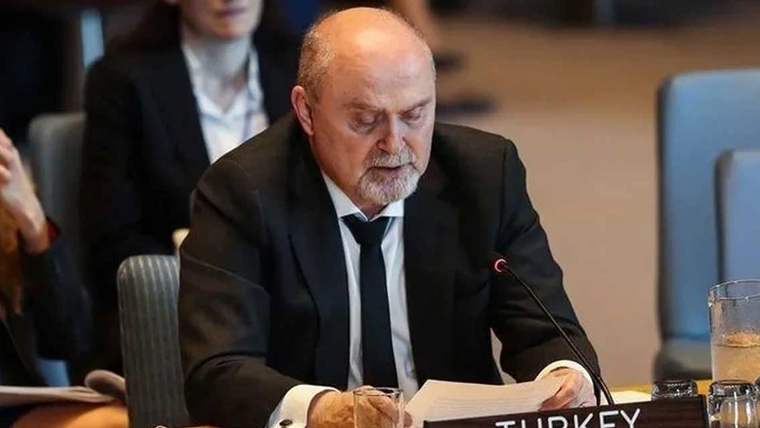 Koltukta oturan bir kişi, belgeleri okurken mikrofonun önünde konuşuyor.