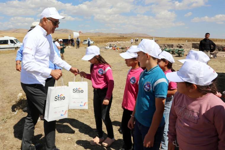 Açık arazide beyaz şapkalı çocuklar, ellerinde STSO logolu çantalar bulunan bir yetişkinin yanında duruyor.