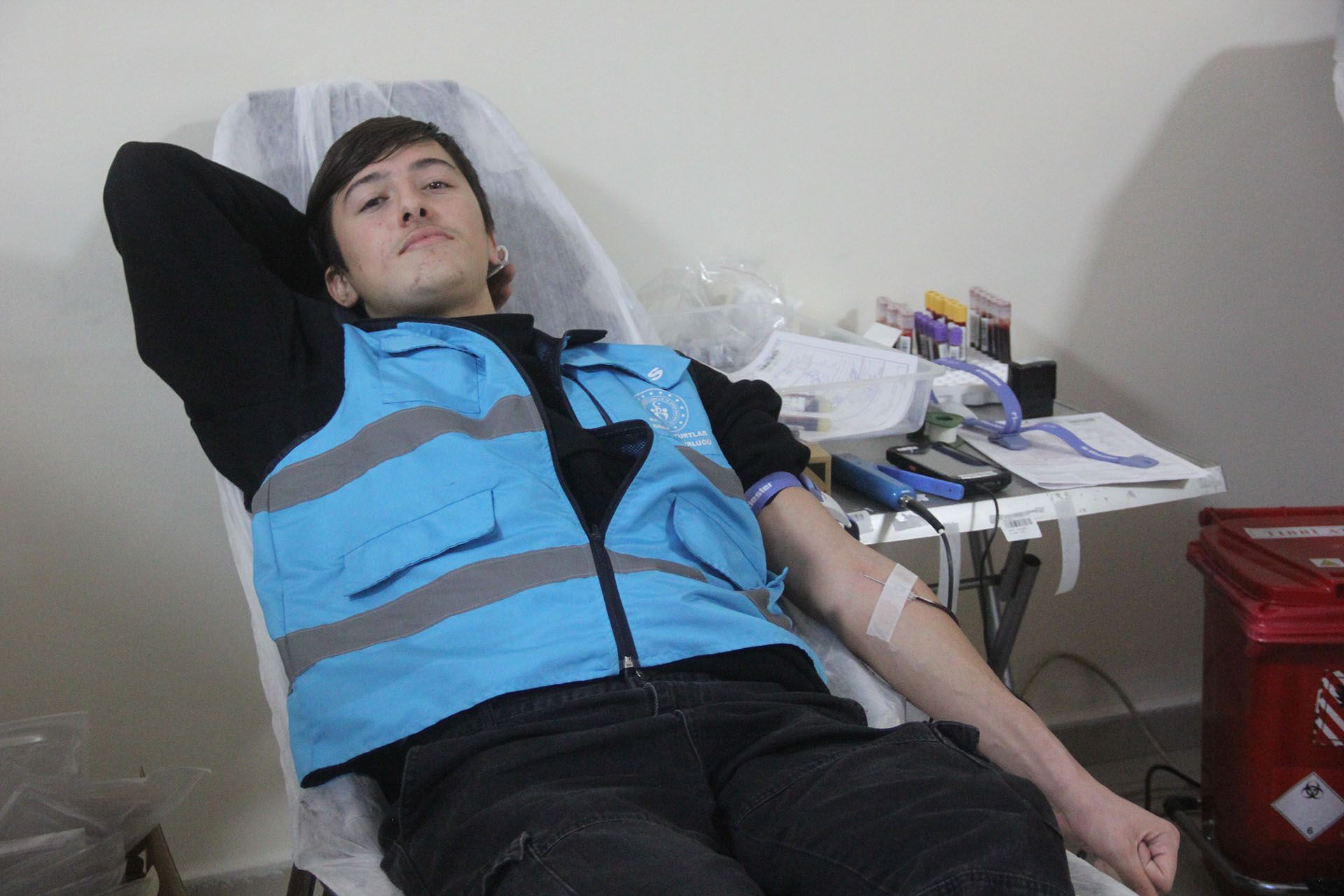 Mavi reflektörlü yelek giyen genç bir kişi, kolunda bantla sağlık yatağında uzanıyor.