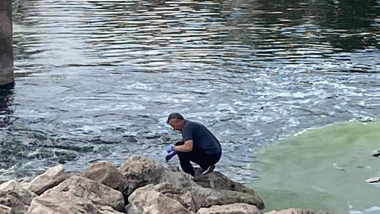 Kayalık kıyıda çömelmiş bir kişi, elindeki mavi nesneyle akıntılı ve bulanık suya bakıyor.
