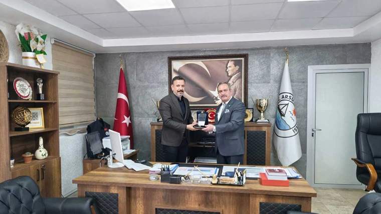 İki kişi, bir masa etrafında durarak plaket takdim ediyor. Arka planda Türk bayrağı ve Arsuz Belediyesi logosu var.