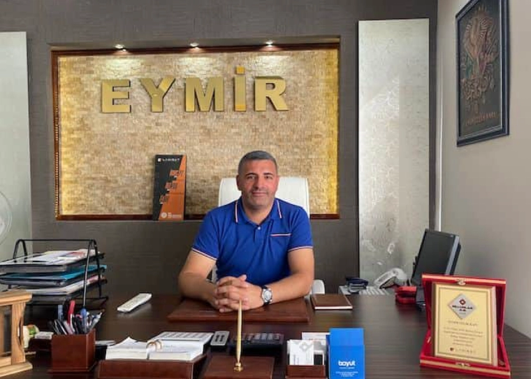 Masasında oturan bir kişi, arkasında “EYMİR” yazılı pano ve çevresindeki ofis eşyalarıyla görülüyor.Eymir Çelik Kapı - Sivas TSO 27 Kalite adayı