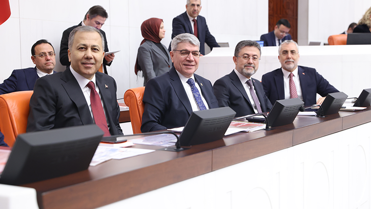 Toplantı salonunda takım elbiseli kişiler, masadaki ekranların önünde sıralı koltuklarda oturuyor.