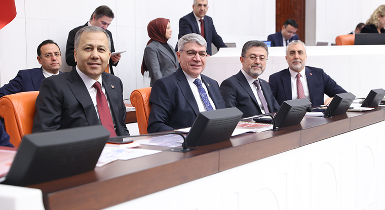 Toplantı salonunda takım elbiseli kişiler, masadaki ekranların önünde sıralı koltuklarda oturuyor.