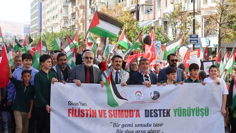 Cadde üzerinde Filistin bayrakları taşıyan kalabalık, “Filistin ve Sumud’a destek yürüyüşü” yazılı pankartı tutuyor.