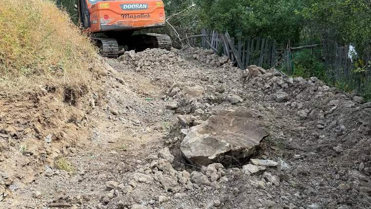Turuncu paletli iş makinesi, ağaçlık alanda taş ve toprakla kaplı bir yol üzerinde duruyor.