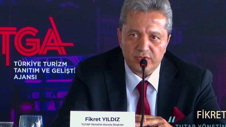 Mikrofonun önünde konuşan bir kişi, masanın üzerinde ismi yazılı bir kart var.