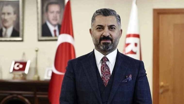 Takım elbiseli sakallı bir erkek, Türk bayrakları ve duvardaki portrelerin önünde kameraya bakıyor.