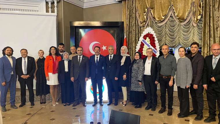 Bir grup insan, arka planda Türk bayrağı ve çiçek aranjmanı ile birlikte poz veriyor.