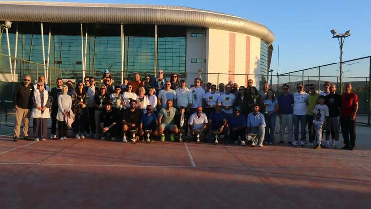 Tenis kortunda, spor kıyafetleri giyen kalabalık bir grup, cam cepheli spor binasının önünde birlikte duruyor.