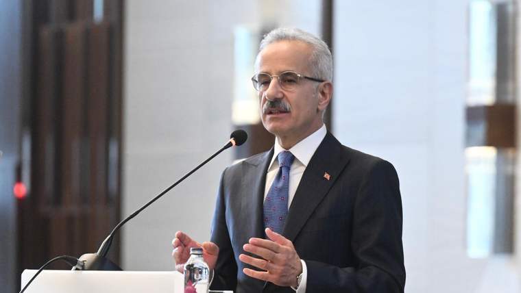Kürsüde konuşan gözlüklü bir kişi, takım elbisesiyle el hareketi yaparken önünde mikrofon ve su şişesi bulunuyor.
