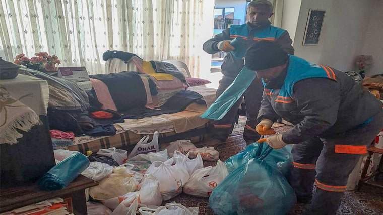 Dağınık bir oturma odasında iki kişi, eldivenlerle yerdeki dolu çöp poşetlerini topluyor.