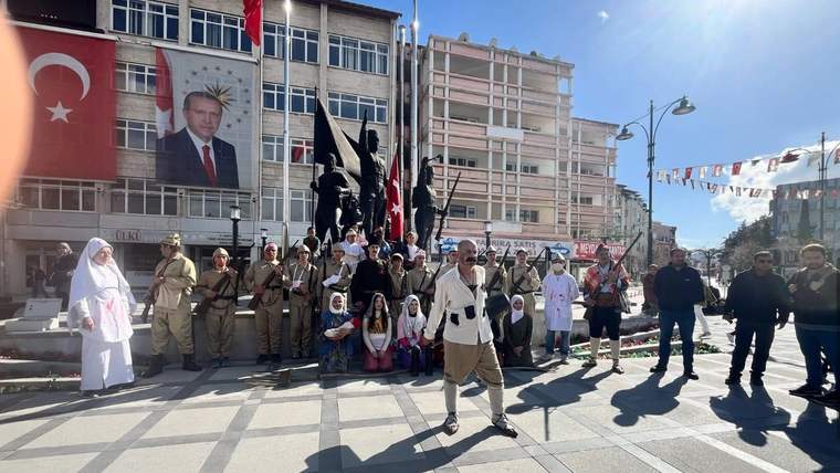 Meydanda tarihi kıyafetler giyen kişiler, Türk bayrakları ve flamalar önünde yan yana duruyor.