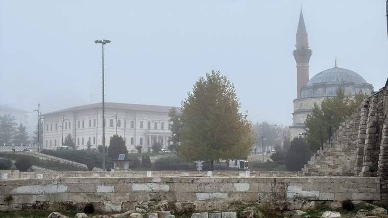 Sisli havada kubbeli ve minareli bir cami, büyük bir yapı ve taş duvarın ardında görülüyor.