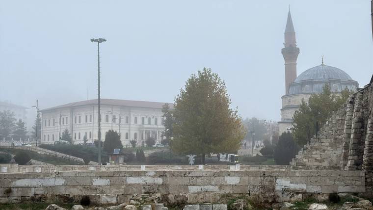 Sisli havada kubbeli ve minareli bir cami, büyük bir yapı ve taş duvarın ardında görülüyor.