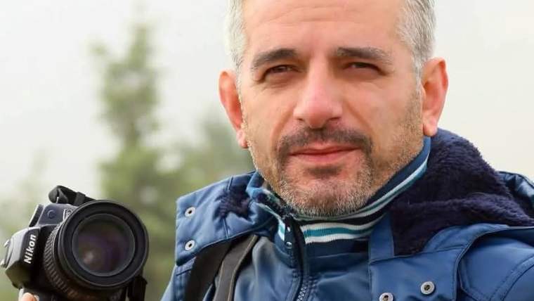 Kamerayı elinde tutan bir erkek, mavi mont giymekte ve hafif gülümsemekte.