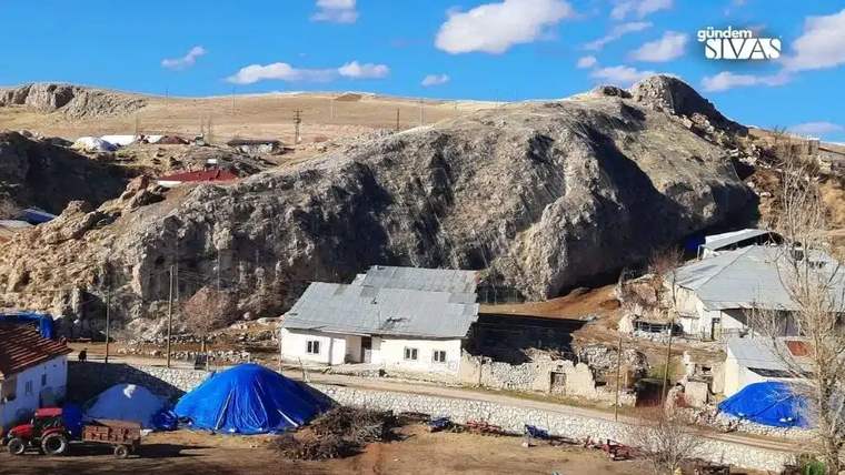 Kayalık yamaç önündeki köyde tek katlı evler, mavi brandalar ve bir traktör görülüyor.