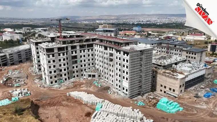 İnşaat halindeki bir hastane binası, çevresinde inşaat malzemeleri ve iş makineleri var.