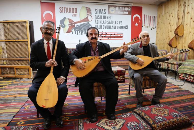 Üç kişi, halılarla döşeli odada bağlama çalıyor; arkalarında dernek adının yazılı olduğu pankart bulunuyor.