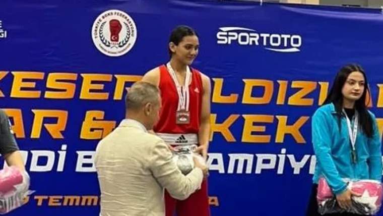 Boks ringinde madalya alan genç bir kadın, ödülünü alırken gülümsüyor.