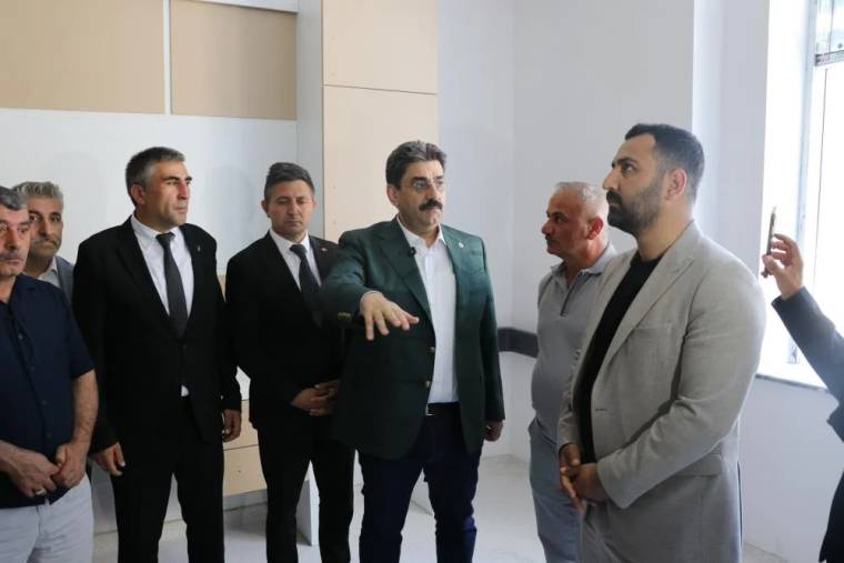 Aydınlık bir iç mekânda duran bir grup erkekten biri elini uzatırken diğerleri onu dinliyor.