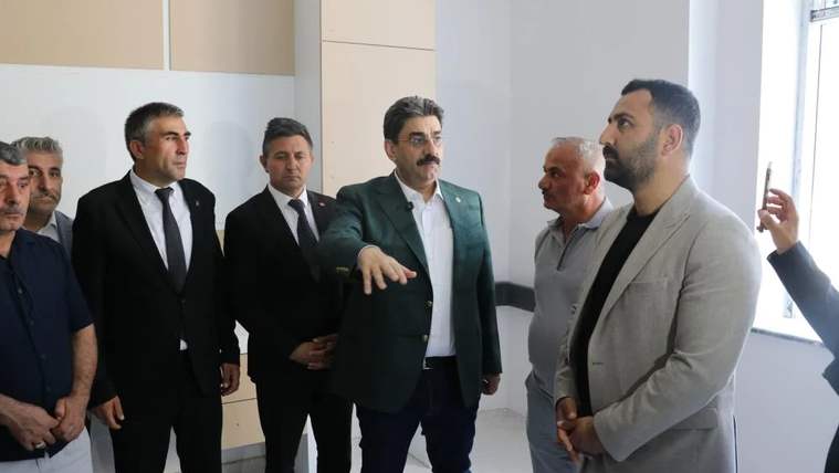 Aydınlık bir iç mekânda duran bir grup erkekten biri elini uzatırken diğerleri onu dinliyor.