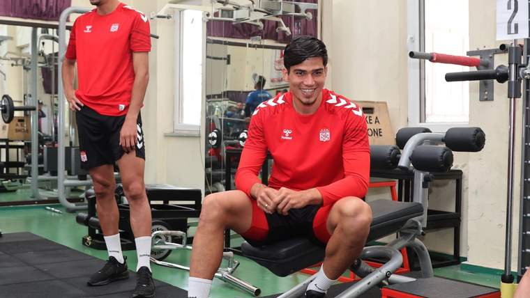 Spor salonunda kırmızı formalar giyen iki futbolcu, ağırlık ve egzersiz aletleri arasında görülüyor.