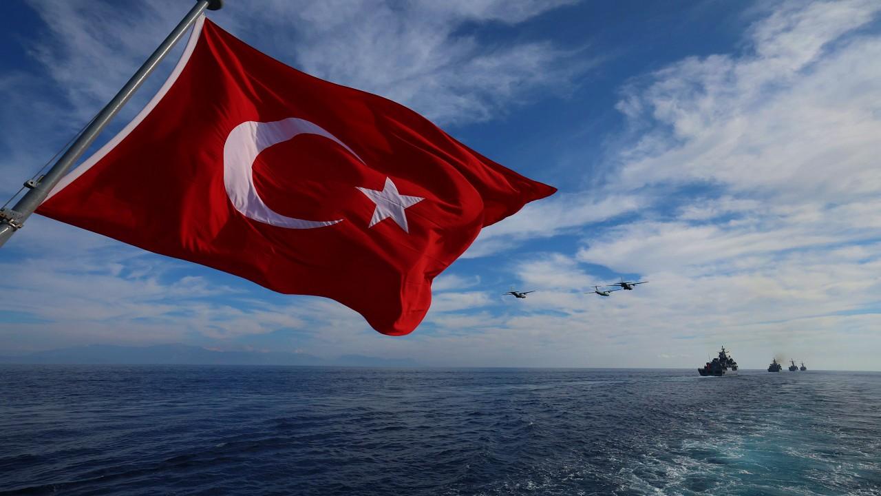 Deniz üzerinde dalgalanan Türk bayrağının arkasında uçaklar ve ufukta birkaç gemi görülüyor.
