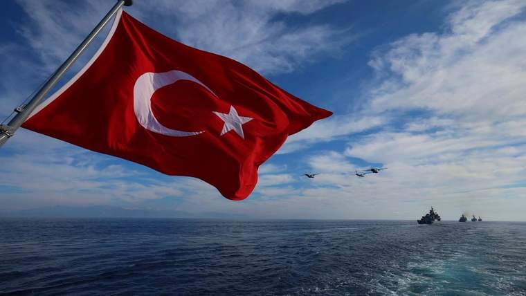 Deniz üzerinde dalgalanan Türk bayrağının arkasında uçaklar ve ufukta birkaç gemi görülüyor.
