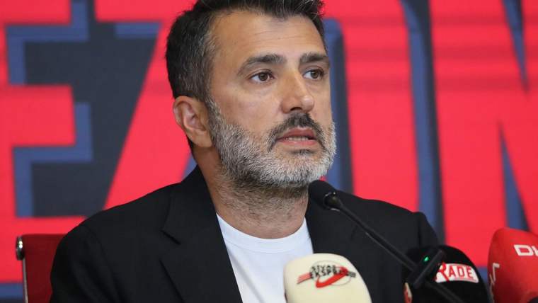 Mikrofonlar önünde konuşan sakallı bir kişi, siyah ceket ve beyaz tişört giyiyor; arka planda kırmızı pano bulunuyor.