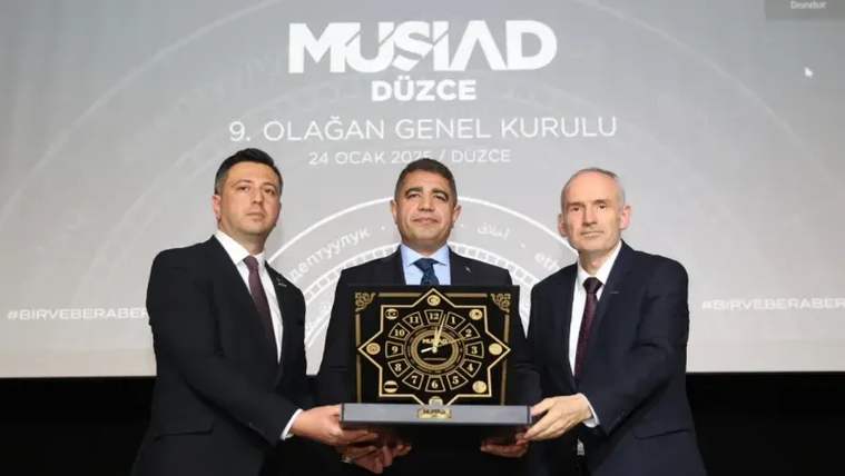 Üç takım elbiseli kişi, üzerinde saat motifleri bulunan plaketi sahnede birlikte tutuyor; arkada MÜSİAD Düzce yazısı görülüyor.