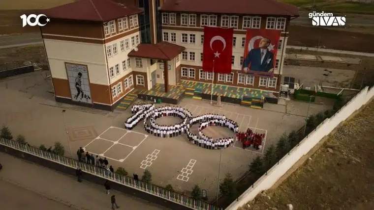 Öğrenciler, okul bahçesinde 100 sayısını oluşturmuş. Arkada Türk bayrağı ve Atatürk posteri görünmekte.