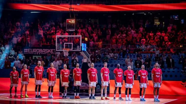 Basketbol sahasında kırmızı formalı oyuncular sıralanırken tribünlerde kalabalık ve üstte Türk bayrağı görülüyor.