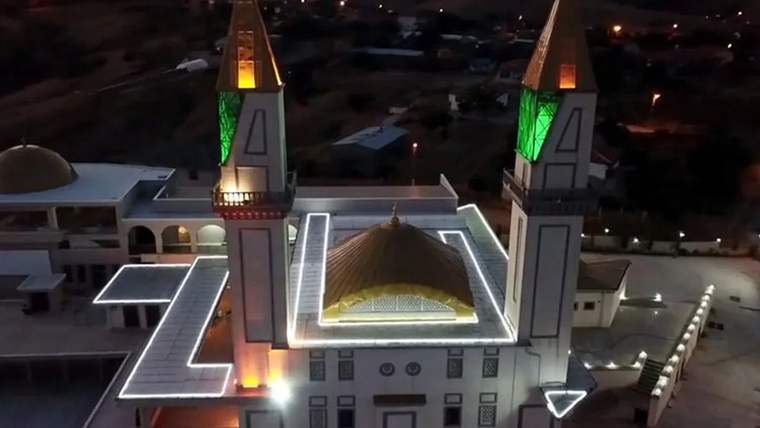 Işıklandırılmış bir cami binası, iki minaresi ve altın kubbesiyle gece görünümünde.