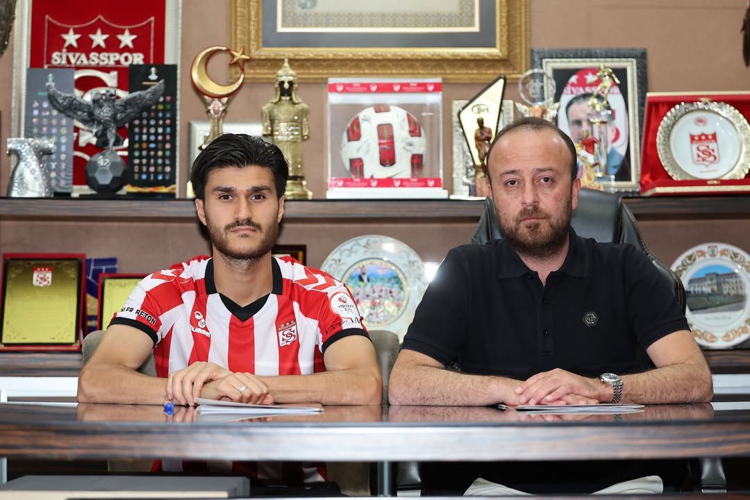 Sivasspor forması giyen bir kişi ile siyah tişörtlü bir kişi, kupa ve plaketlerin önünde masada oturuyor.