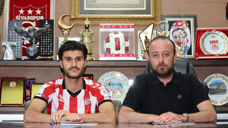 Sivasspor forması giyen bir kişi ile siyah tişörtlü bir kişi, kupa ve plaketlerin önünde masada oturuyor.