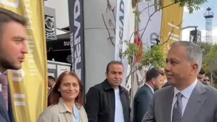 Gri takım elbiseli bir kişi, sarı pankartların bulunduğu alanda başka biriyle el sıkışırken görülüyor.