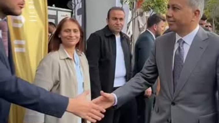 Gri takım elbiseli bir kişi, sarı pankartların bulunduğu alanda başka biriyle el sıkışırken görülüyor.