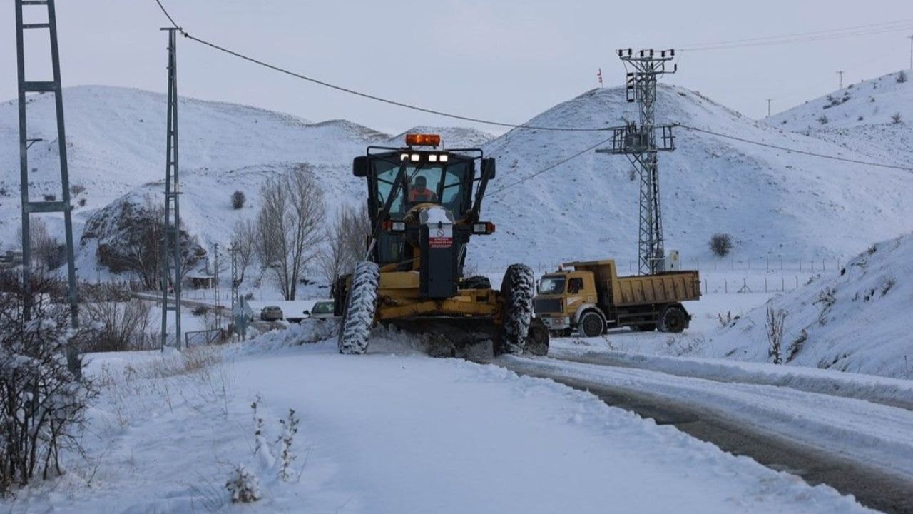 Karlarla kaplı yolda bir iş makinesi ilerlerken arkasında sarı damperli kamyon ve dağlık arazi görülüyor.
